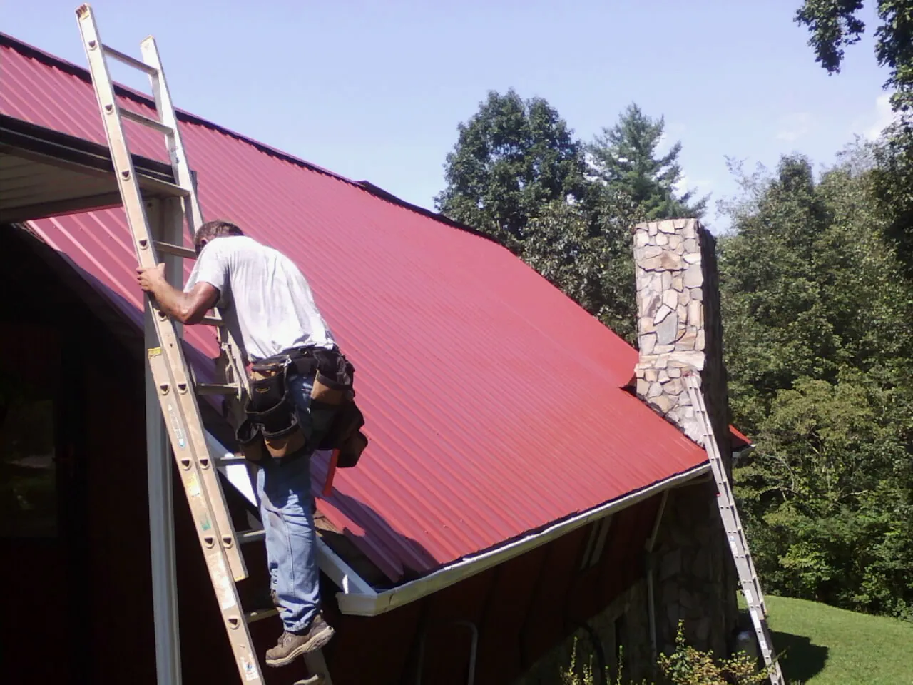 Roofer on a ladder beside a red metal roof and stone chimney — Soffit & Fascia Repair by Roofing Brothers in Mount Kisco
